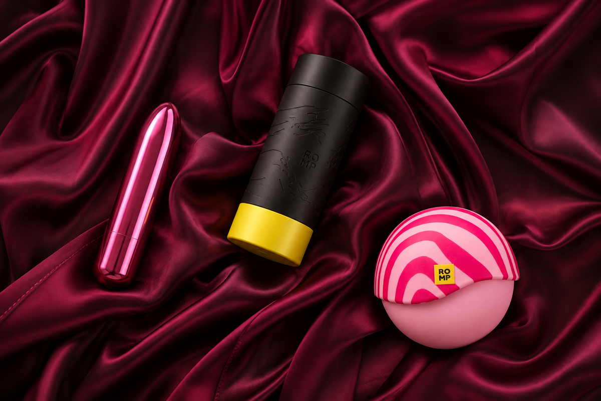 Compact vibrator, manual stroker and water-based lubricant beside a closed gift box on cream and muted-plum fabric